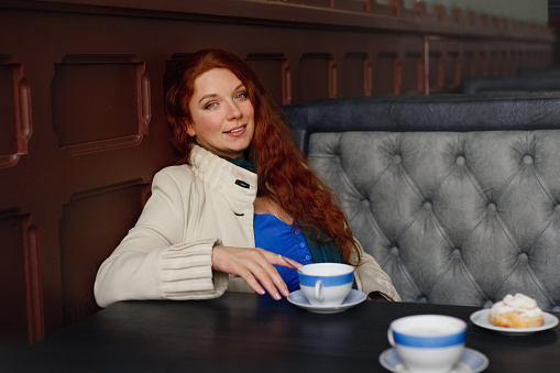 young smiling red-haired European woman in beige coat and blue dress sits in old cafe, lifestyle