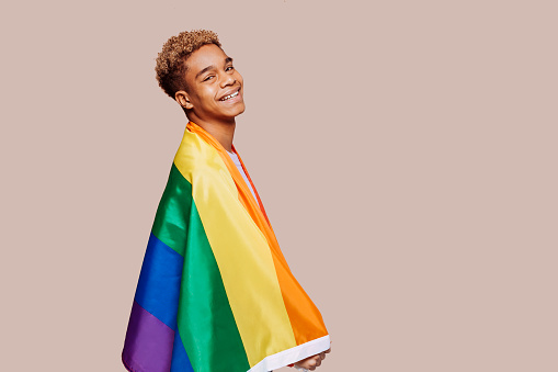 Side view portrait of cheerful young afro latin black man with a gay pride rainbow flag at studio over beige background. Homosexual lgbtiq concept, rainbow flag, celebrating parade. Copy space.