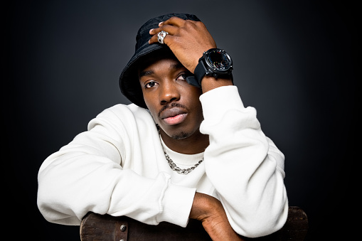 Headshot of young man wearing white sweatshirt, black bucket hat, watch and silver ring and necklace, looking at camera. Studio shot on black background.