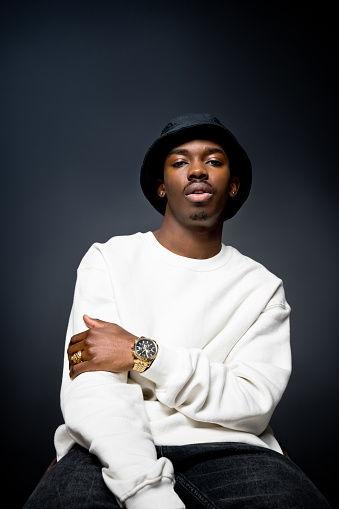 Fashion portrait of young man wearing white sweatshirt, black bucket hat, gold watch and ring, looking at camera. Studio shot on black background.