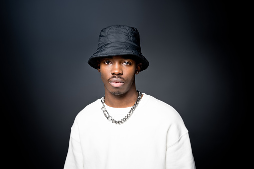 Fashion portrait of young man wearing white sweatshirt, black bucket hat, looking at camera. Studio shot on black background.