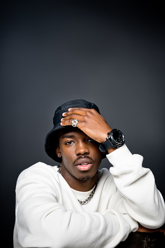 Headshot of young man wearing white sweatshirt, black bucket hat, watch and silver jewelry looking at camera. Studio shot on black background.