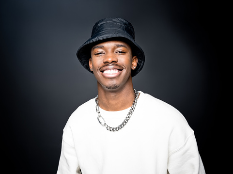 Fashion portrait of cheerful young man wearing white sweatshirt, black bucket hat, smiling at camera. Studio shot on black background.