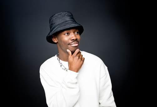 Fashion portrait of smiling young man wearing white sweatshirt, black bucket hat, looking away with hand on chin. Studio shot on black background.