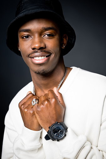 Headshot of happy young man wearing white sweatshirt, black bucket hat, watch and silver ring, smiling at camera. Studio shot on black background.