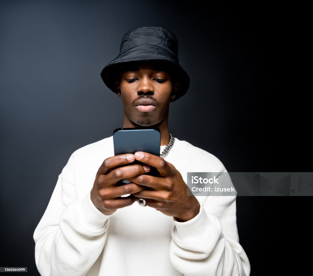 Young man using smart phone Portrait of handsome young man wearing white sweatshirt and black bucket hat, using mobile phone. Studio shot on black background. Focus on hands. Using Phone Stock Photo