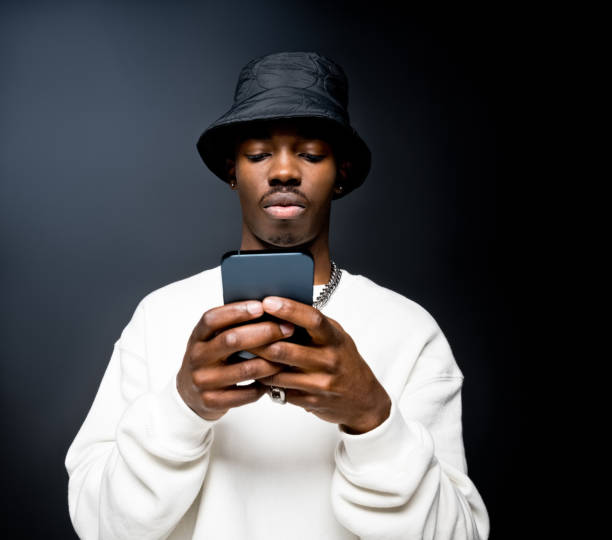 Young man using smart phone Portrait of handsome young man wearing white sweatshirt and black bucket hat, using mobile phone. Studio shot on black background. Focus on hands. rich black men pictures stock pictures, royalty-free photos & images