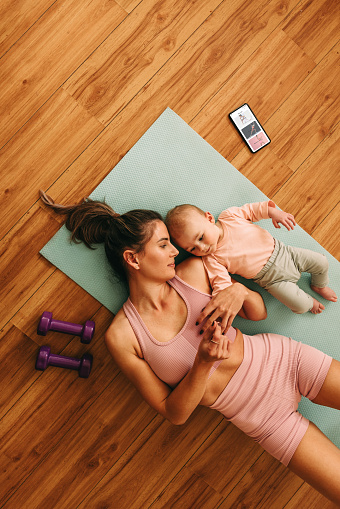 Top view of a young mom lying on an exercise mat with her baby. Mother and baby taking a break from working out. New mom bonding with her baby during her post-natal fitness routine.