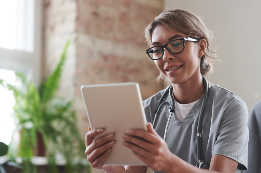 Portrait of attractive female doctor wearing eyeglasses sitting at desk holding digital tablet talking to patient on video call