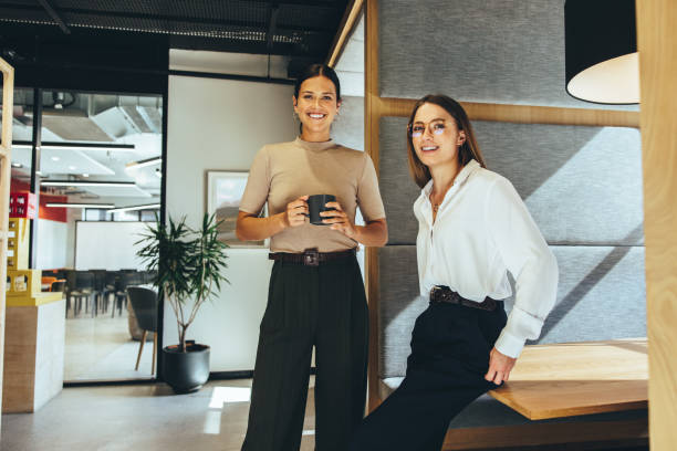 Female entrepreneurs smiling at the camera Cheerful female entrepreneurs smiling at the camera while standing in a modern co-working space. Two happy young businesswomen taking a coffee break while working together. open plan stock pictures, royalty-free photos & images