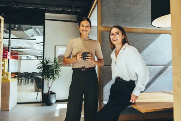 Cheerful female entrepreneurs smiling at the camera while standing in a modern co-working space. Two happy young businesswomen taking a coffee break while working together.