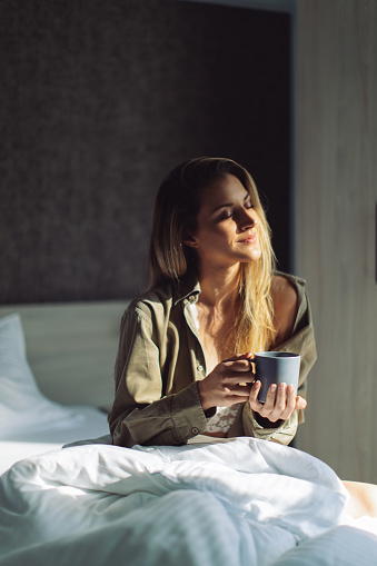 Beautiful young woman sitting on bed in bedroom and enjoying morning sunlight next to the window at her home and drinking coffee. She is very happy and relaxed