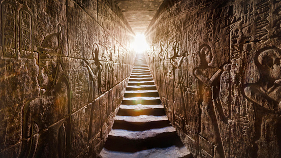 Templo de Edfu, Egipto. Pasaje flanqueado por dos paredes brillantes llenas de jeroglíficos egipcios, iluminados por una cálida luz de fondo naranja de una puerta al final de las escaleras. photo
