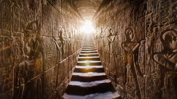 Photo of Temple of Edfu, Egypt. Passage flanked by two glowing walls full of Egyptian hieroglyphs, illuminated by a warm orange backlight from a door at the end of the stairs.