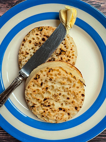 Stock photo showing close-up, elevated view of a blue and white Cornish ware plate containing freshly baked, warm, homemade buttered crumpets with melting butter.