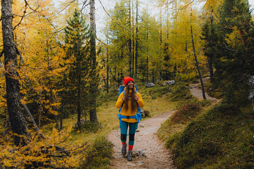 Young woman backpacker in red hat enjoying the hiking trip in the autumn woods, admiring the beautiful mountain peaks during sunny autumn day