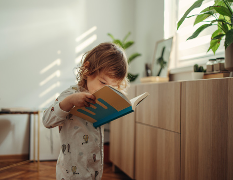 Cute baby girl is standing in her pyjamas holding and looking at the book. There are shadows on the wall from the morning sunlight.
