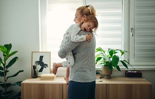 Cute baby girl is yawning on her mom's shoulder in the morning. Her mother is standing and holding her in her arms.