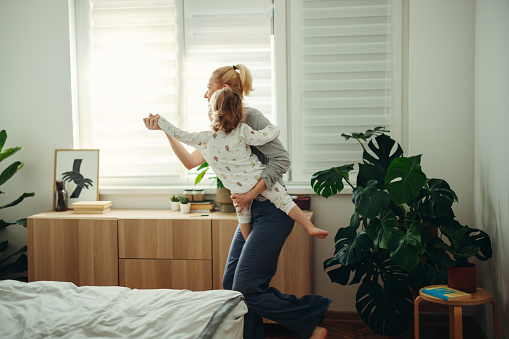 Happy mom is holding her daughter in her arms and dancing with her. There are plants and dimmed light in the room.