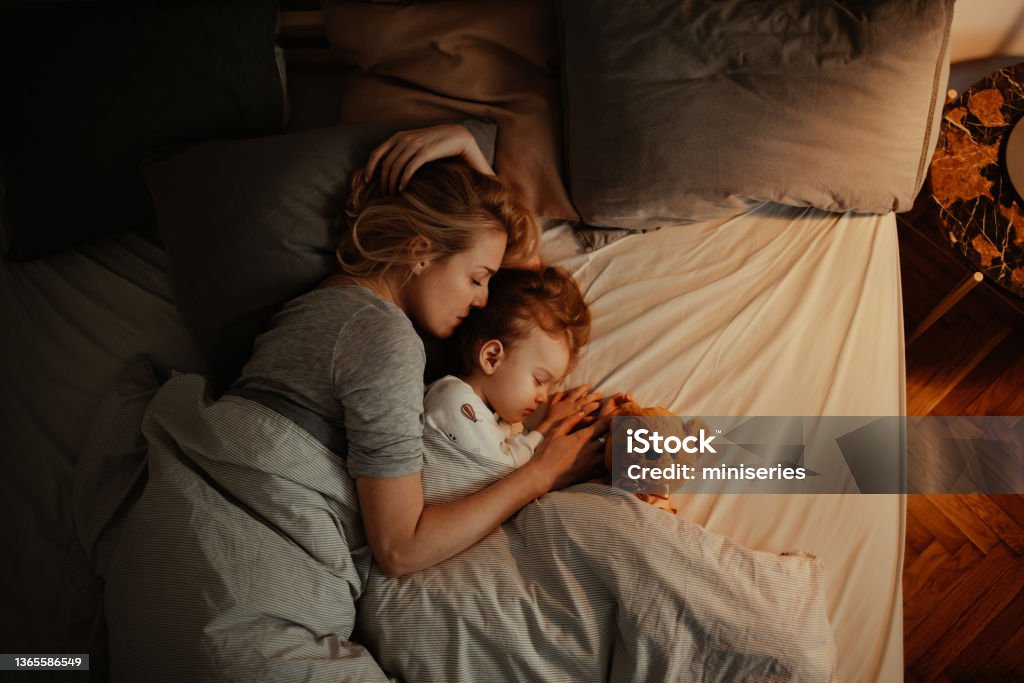 Loving Mother and Daughter Sleeping Together in Bed in the Evening Beautiful mother and daughter sleeping together in bed. It is the evening and there is lamp on. Mom is holding her daughter and keeping her close. There is a teddy bear under the covers. Sleeping Stock Photo