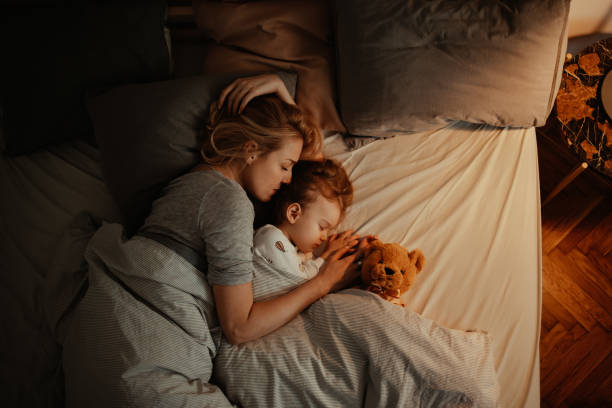 Loving Mother and Daughter Sleeping Together in Bed in the Evening Beautiful mother and daughter sleeping together in bed. It is the evening and there is lamp on. Mom is holding her daughter and keeping her close. There is a teddy bear under the covers. sleeping stock pictures, royalty-free photos & images
