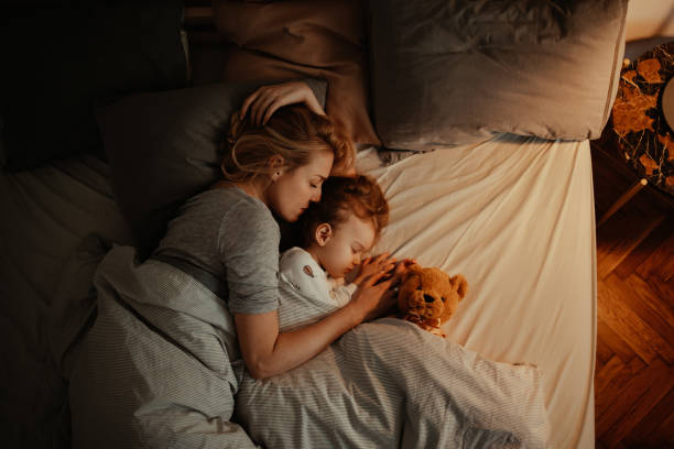Loving Mother and Daughter Sleeping Together in Bed in the Evening Beautiful mother and daughter sleeping together in bed. It is the evening and there is lamp on. Mom is holding her daughter and keeping her close. There is a teddy bear under the covers. sleeping mom stock pictures, royalty-free photos & images