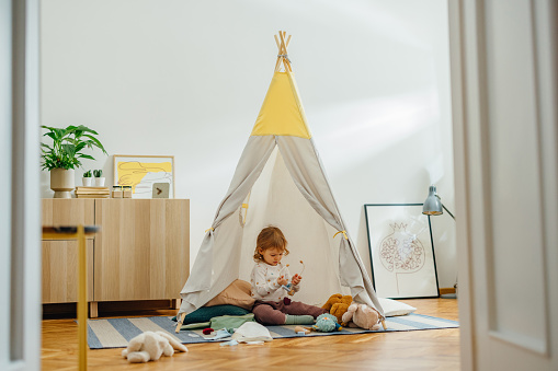 Cute girl and playing in the grey teepee tent. Tent is on the carpet on the floor. There are things and toys around her. She is holding a toy stethoscope.