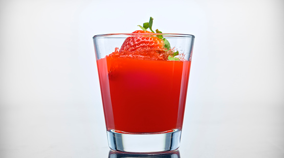 Close-up of fresh strawberry falling into a glass of red juice against white background.
