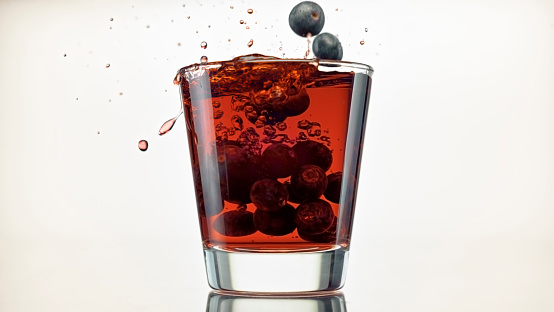 Close-up of fresh blueberries falling into a glass of red juice against white background.