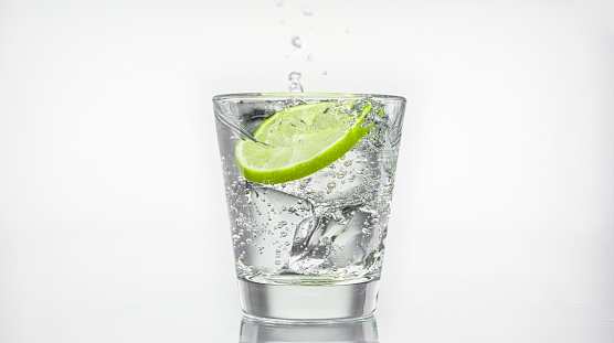 Close-up of slice of lemon falling into a glass of sparkling water with ice against white background.