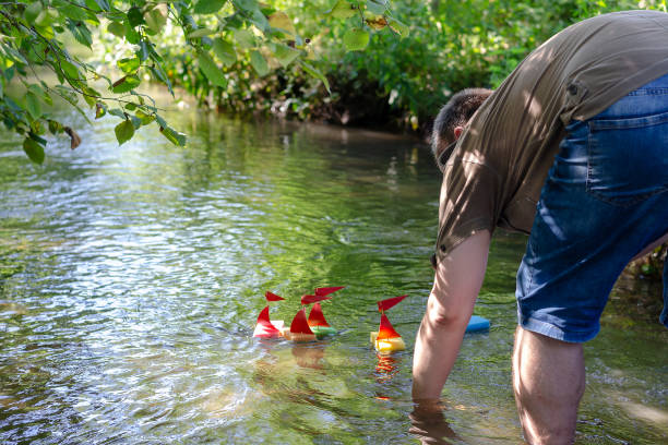 An adult man is launching children's ships while standing in the river. Person bent down in the water makes homemade boats. Toy ships made of kitchen sponges with red sails and flagpoles.