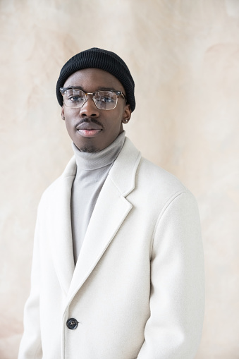 Fashion portrait of handsome young man wearing white coat, turtleneck and black beanie knit hat looking at camera. Studio shot on beige background.