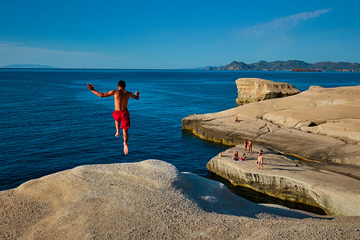 Sarakiniko, Greece - May 23, 2019: Young tourists jump off the white rocks cliff on famous tourist attraction of Milos island Sarakiniko beach and dive into Aegean sea, Milos island , Greece