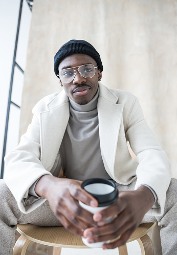 Fashion portrait of handsome young man wearing white coat, turtleneck and black beanie knit hat, sitting on chair in studio and holding cup of coffee. Studio shot on beige background.
