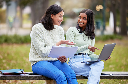 Smiling teen boy explaining homework over book to friend while sitting on railing against trees in campus