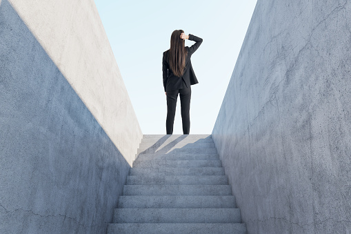 Back view of thoughtful businesswoman standing on top of abstract concrete stairs to success with sunlight. Growth and leadership concept
