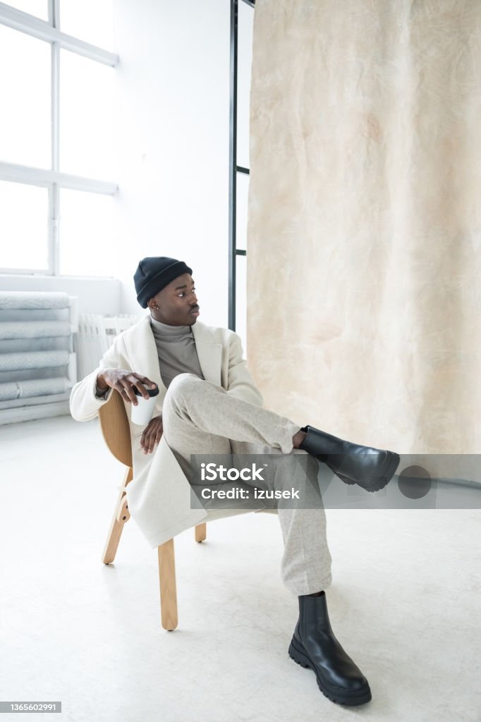 Elegant young man wearing white winter coat Fashion portrait of handsome young man wearing white coat, turtleneck and black beanie knit hat and boots sitting on chair in studio next to beige background. Fashion Stock Photo
