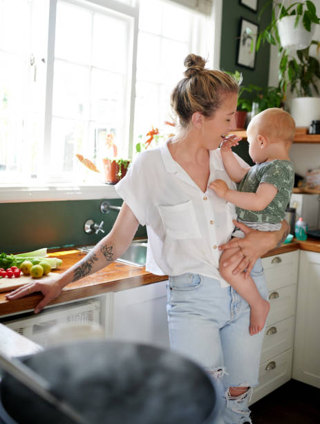 foto de una madre de pie y cargando a su hijo mientras preparaba comida en la cocina - playing playful baby contemporary fotografías e imágenes de stock