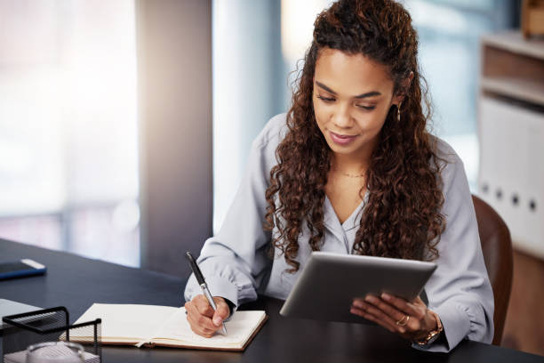 istock Shot of a young businesswoman using her digital tablet while writing in her diary 1365606643