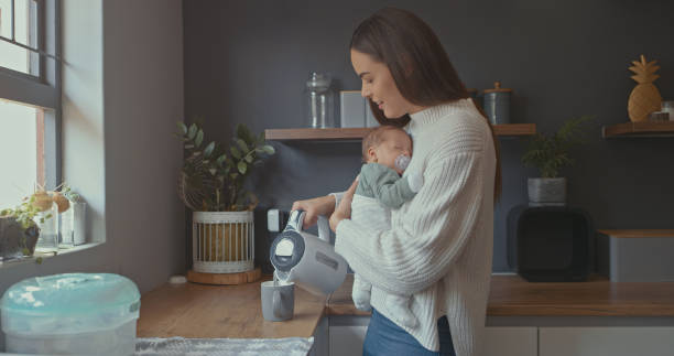 Shot of a young woman pouring herself a cup of tea while holding her baby stock photo