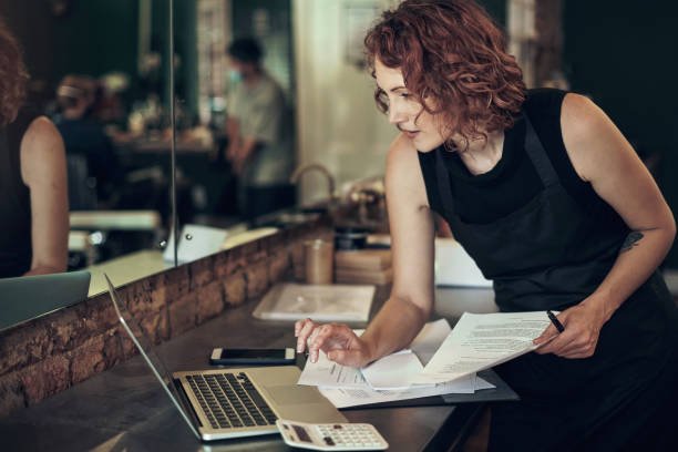 photo d’une jeune coiffeuse séduisante debout et utilisant son ordinateur portable tout en calculant ses finances dans son salon - document strategy insurance business photos et images de collection