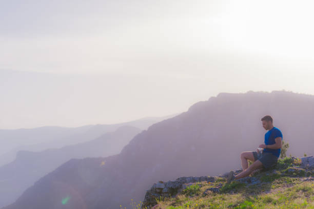 istock Handsome athlete sitting at a rocky peak while looking at the breathtaking mountain line and a beautiful lake while wearing a blue shirt and grey shorts. 1365616751