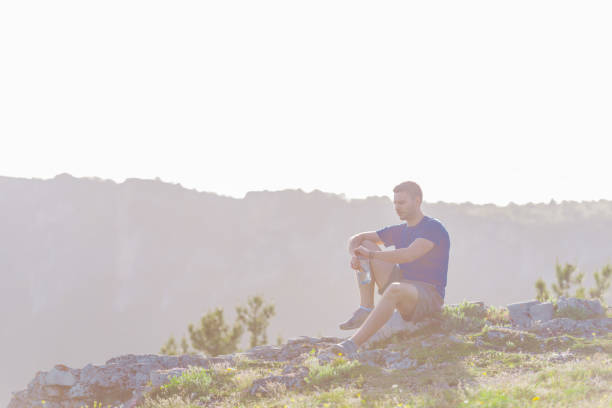 istock Handsome athlete sitting at a rocky peak while looking at the breathtaking mountain line and a beautiful lake while wearing a blue shirt and grey shorts. 1365616752