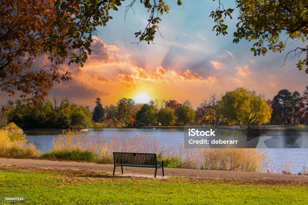 Empty park bench sits by the water with the light of sunset in the background Empty park bench sits by the water with the colorful light of sunset in the background Autumn Stock Photo
