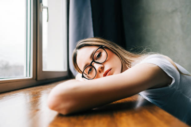 junge gelangweilte millennial-frau auf der fensterbank allein zu hause. müdigkeit oder emotional erschöpftes konzept - human eye rubbing tired glasses stock-fotos und bilder