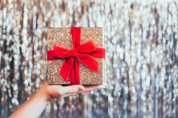 mão feminina segurando caixa de presente dourado brilhante com fita vermelha de veludo com um arco no fundo de lata de ouro brilhante. o conceito de presente surpresa de aniversário, natal. fundo festivo. - bow christmas red velvet - fotografias e filmes do acervo