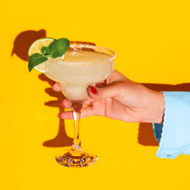 Female hand holding glass with margarita cocktail isolated on bright yellow neon background with shadow. Concept of taste, alcoholic drinks Sweet and sour. Female hand holding glass with margarita cocktail isolated on bright yellow neon background with shadow. Complementary colors, white, blue and yellow. Copy space for ad. Pop art margarita stock pictures, royalty-free photos & images