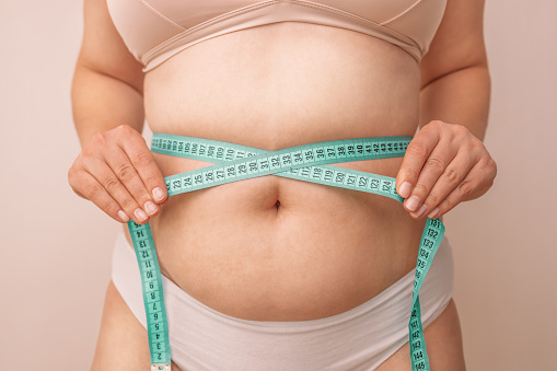 Vientre gordo. Chica caucásica gorda midiendo cintura con cinta métrica verde sobre fondo beige, reduciendo el exceso de peso. Estilo de vida saludable. photo