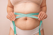 Fat belly. Fat caucasian girl measuring waist with green measuring tape on a beige background, reducing excess weight. Healthy lifestyle.