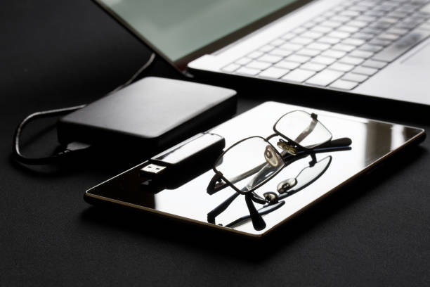An open slim laptop sits next to a tablet, external hard drive, USB stick, and glasses. Backlighting. The concept of data security, workplace, remote and office work. Close-up